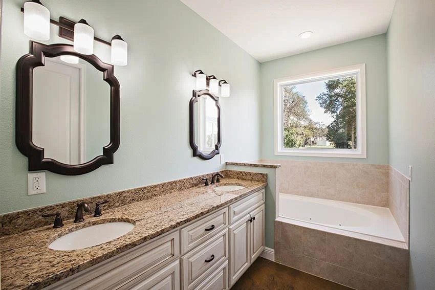 Bathroom with a white vanity, granite top, framed mirrors and a tiled drop-in tub under a window