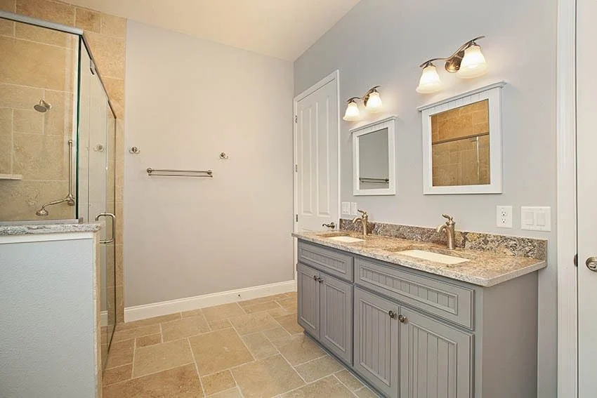 Bathroom with a gray double vanity, granite top, framed mirrors and a glass walk-in shower with travertine tile