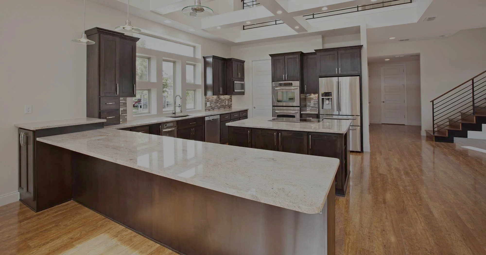 Wide view of a kitchen with dark cabinets, light granite counters, a large island and a coffered ceiling