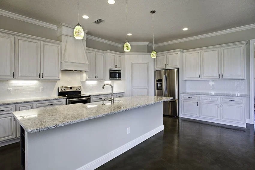 White kitchen with a long granite island, pendant lights, a white range hood and stainless appliances